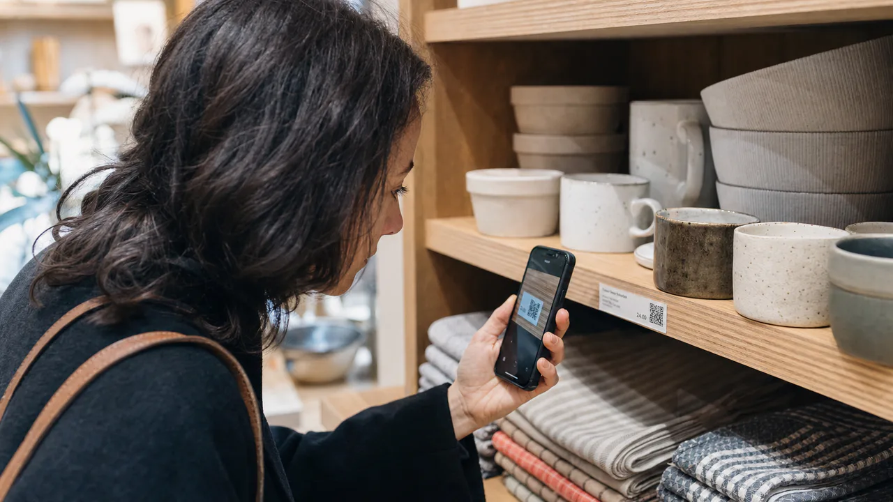 A shopper scans a QR code on a shelf label beside ceramics and folded textiles in a homewares store.